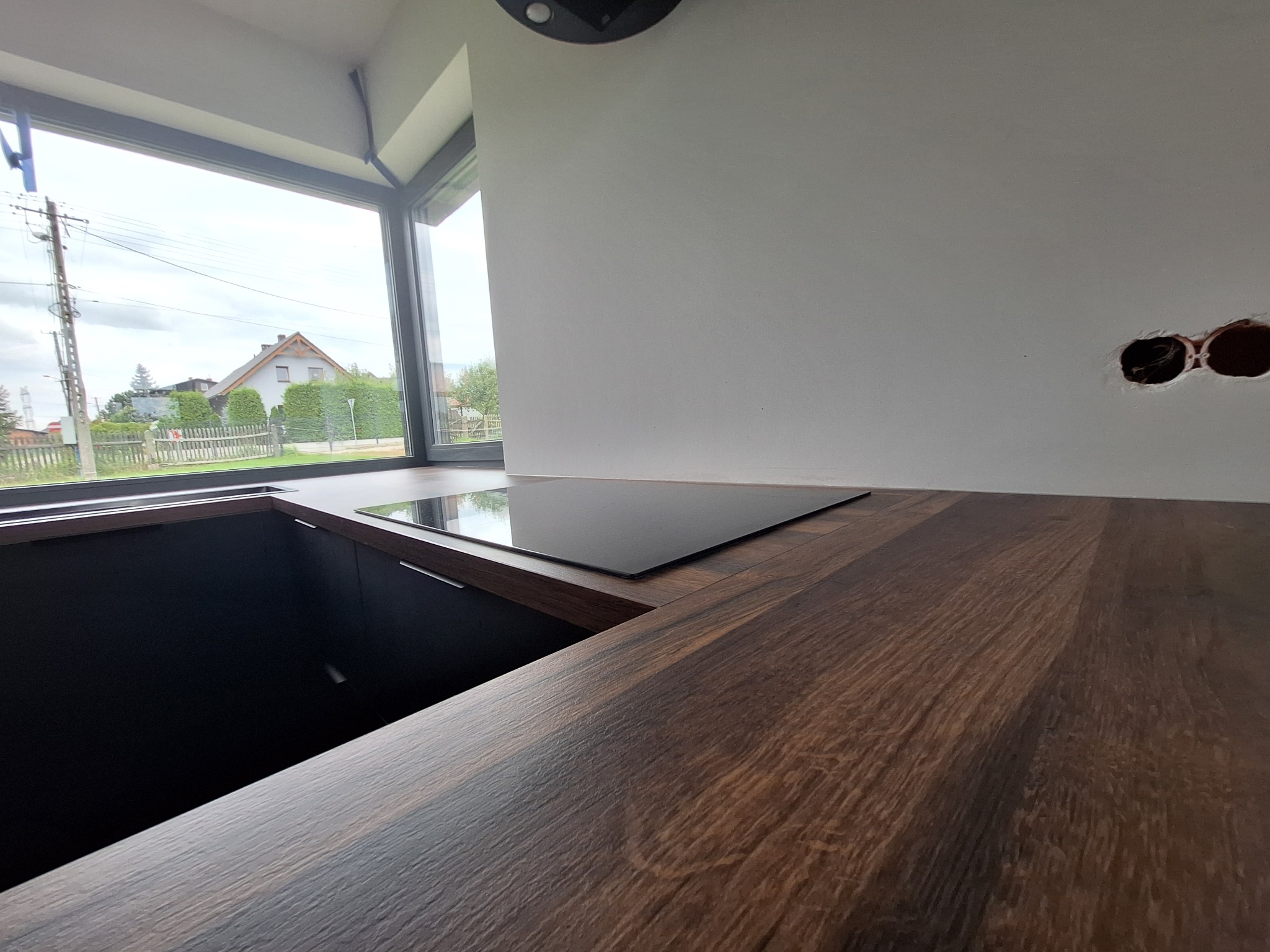 Fragment blatu z ciemnego drewna z wbudowaną płytą grzewczą, widoczne otwory w ścianie na gniazdka elektryczne i okno z widokiem na dom.