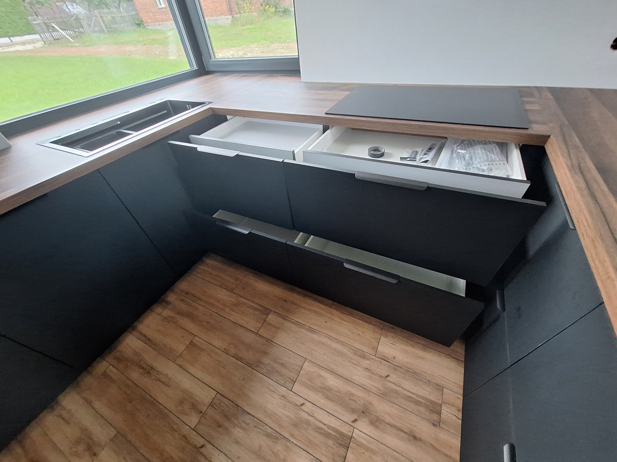 Ciemna, nowoczesna zabudowa kuchenna z drewnianym blatem, widoczne otwarte szuflady z akcesoriami montażowymi, podłoga z paneli imitujących drewno.