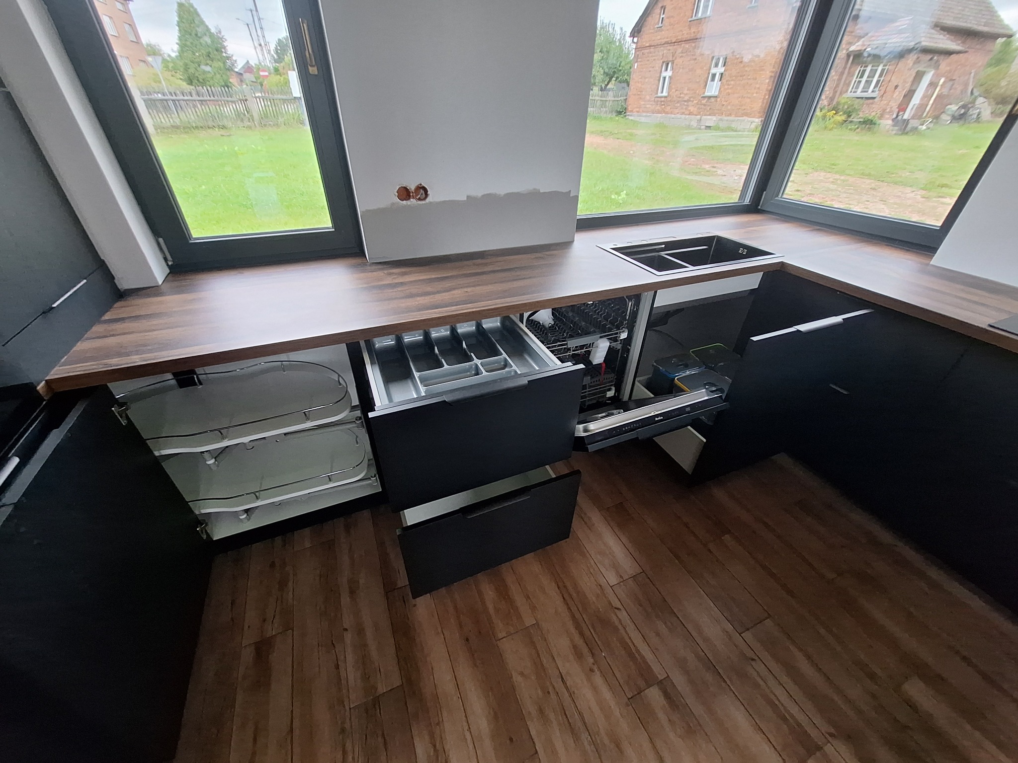 Częściowo zamontowana, nowoczesna kuchnia z czarnymi szafkami, drewnianym blatem i widokiem na ogród przez duże okno. Otwarta szuflada na sztućce i widoczna zmywarka w trakcie montażu.