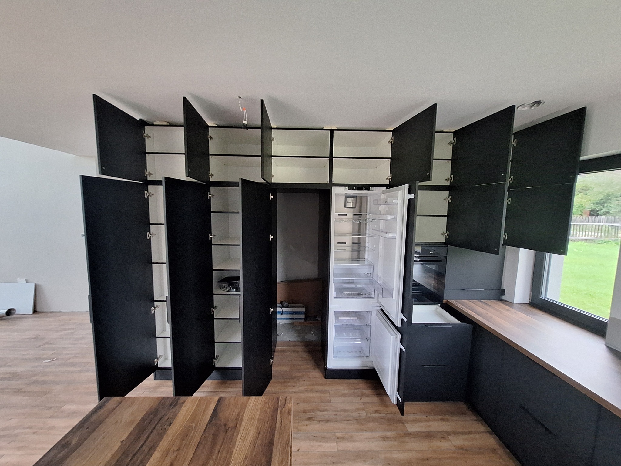 Nowoczesna kuchnia z czarnymi szafkami z otwartymi drzwiczkami, białe wnętrze szafek, lodówka wolnostojąca, drewniany blat i fragment stołu z drewnianym blatem, widoczne elementy instalacji...