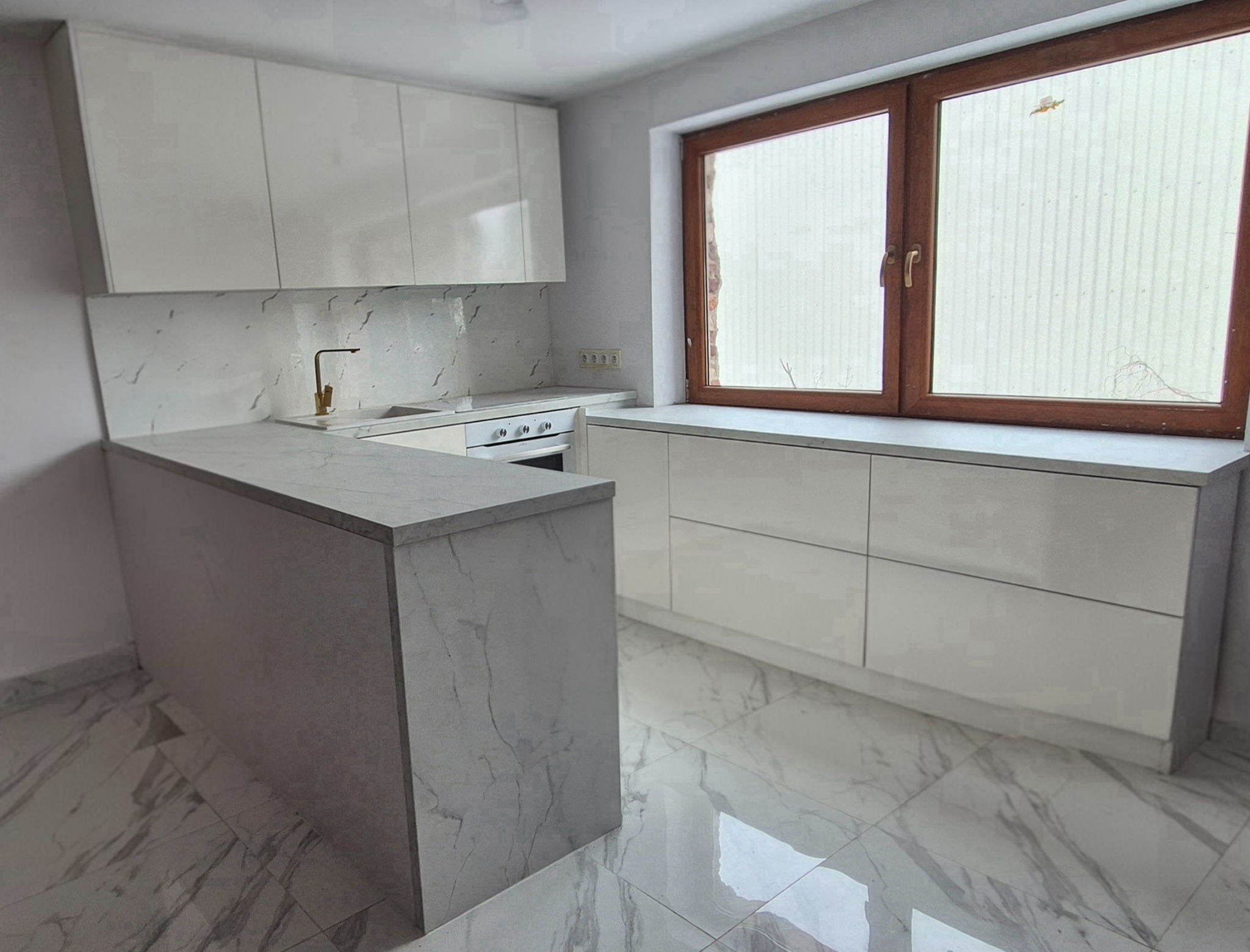 Biała kuchnia z połyskiem, marmurowy blat i backsplash, złota bateria, drewniane okno. Nowoczesny design z wyspą kuchenną i marmurową podłogą.