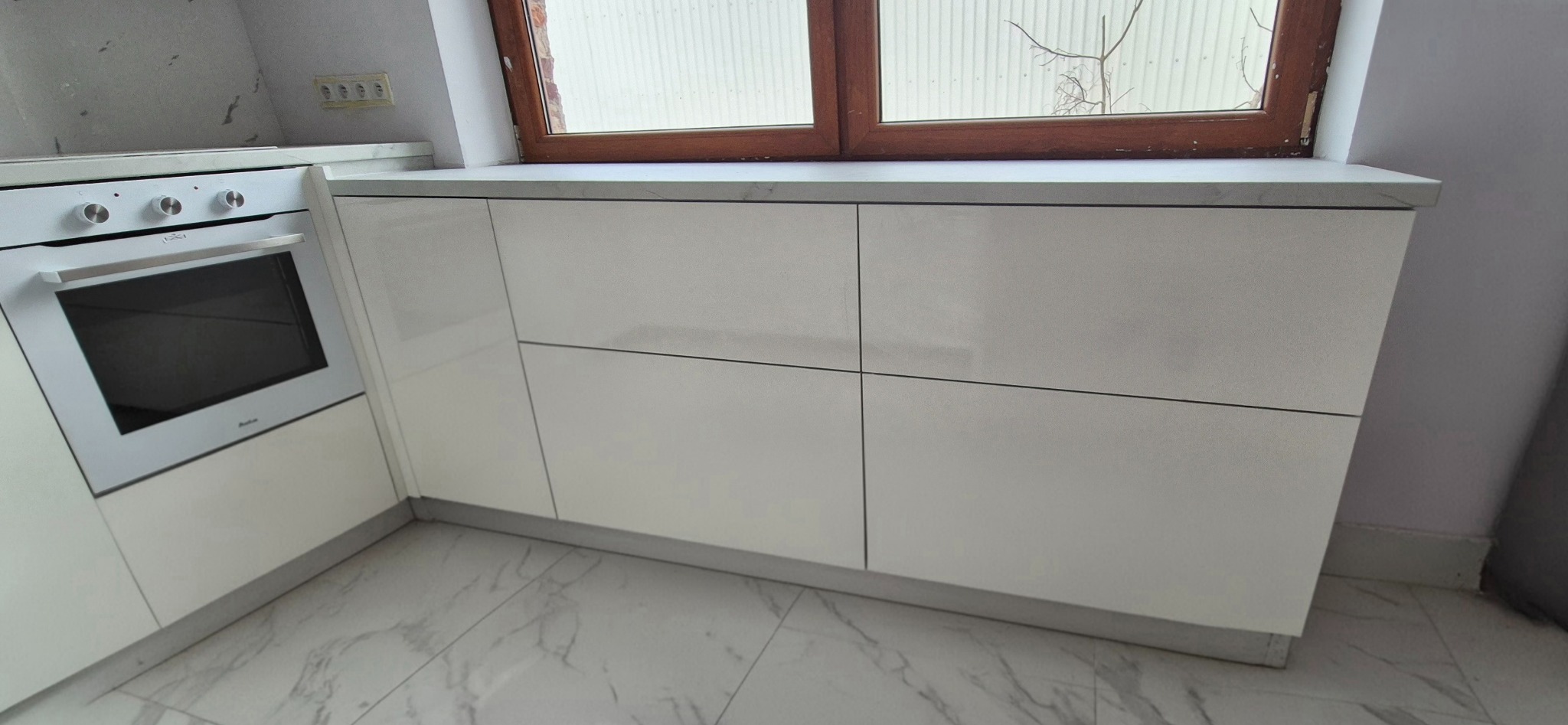 Biała zabudowa kuchenna z piekarnikiem pod oknem, minimalistyczny design, marmurowa podłoga i blat, widoczne gniazdka elektryczne na ścianie.