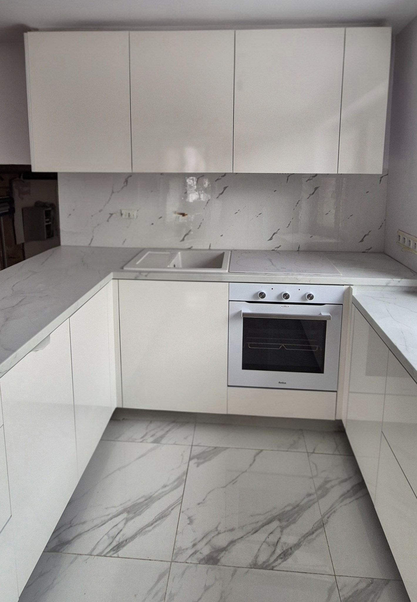 Biała, nowoczesna kuchnia z marmurowym blatem i płytkami, zabudowana pod sufit, z piekarnikiem Amica i płytą grzewczą. Minimalistyczny design.
