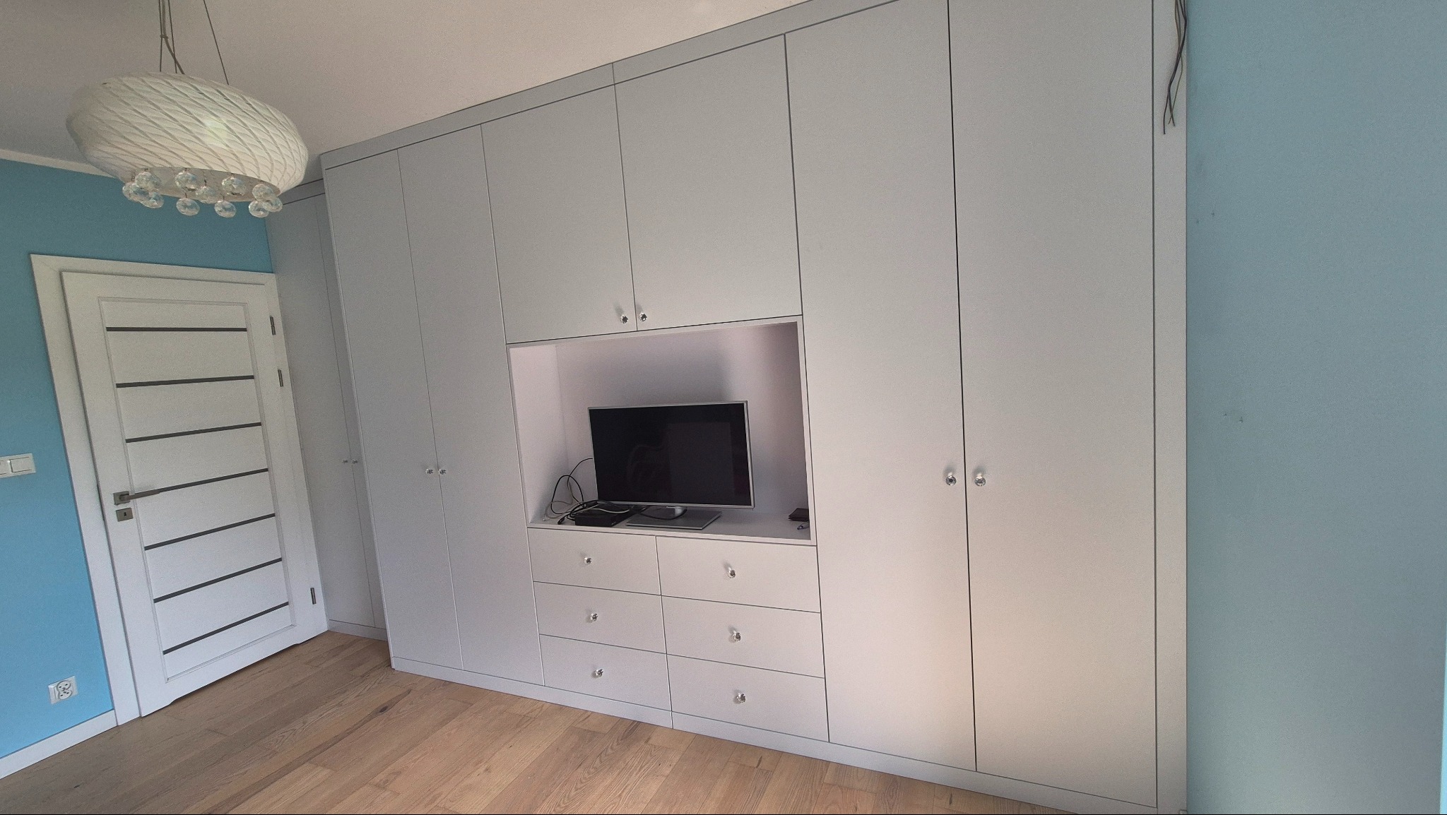 Jasnoszara szafa wnękowa z wbudowanym miejscem na telewizor i szufladami, okrągłe uchwyty, parkiet na podłodze, niebieska ściana w tle.