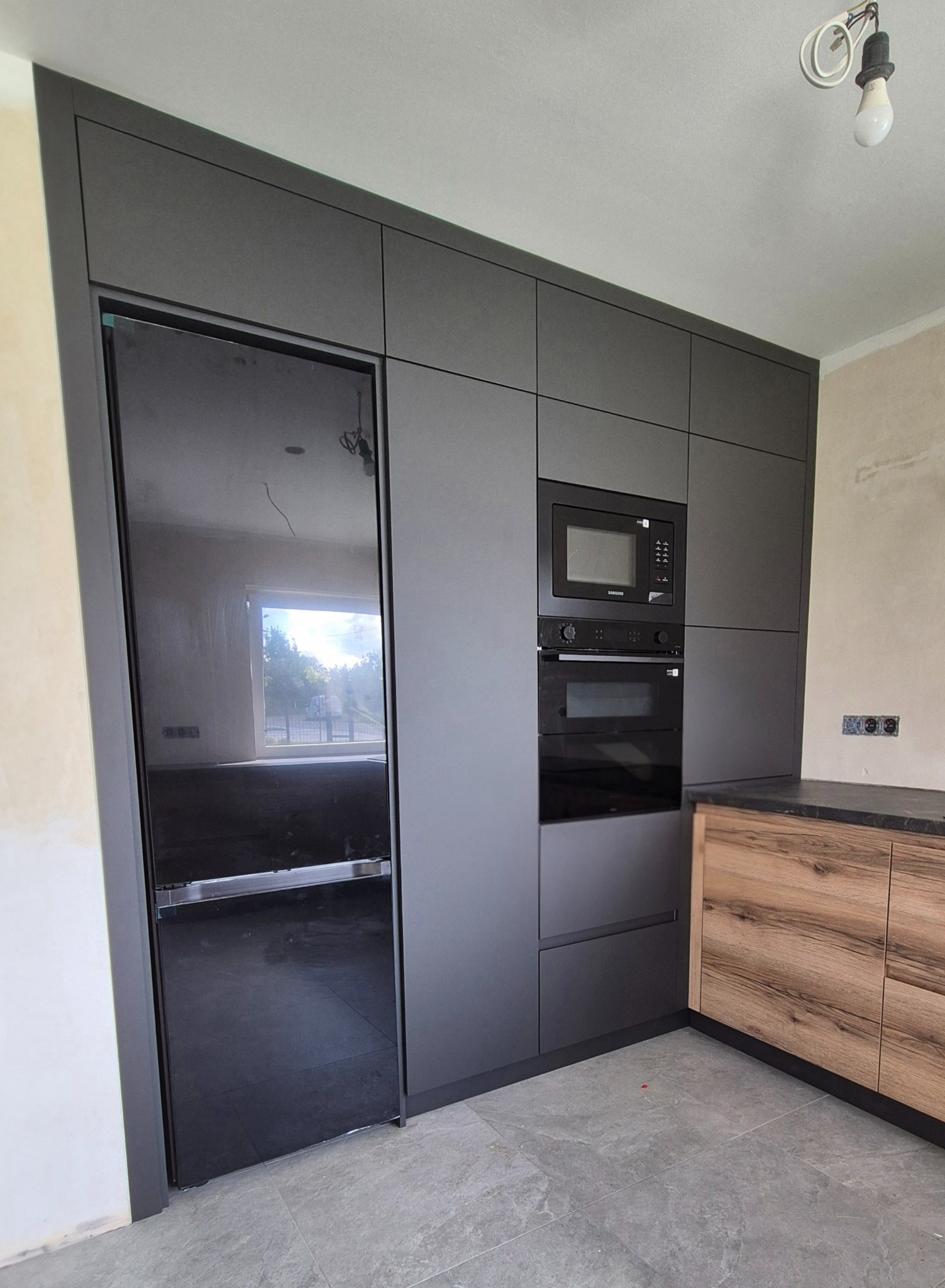 Nowoczesna kuchnia na wymiar w odcieniach szarości z czarnym sprzętem AGD i drewnianymi akcentami. Minimalistyczny design, widoczne niedokończone wykończenie ścian.