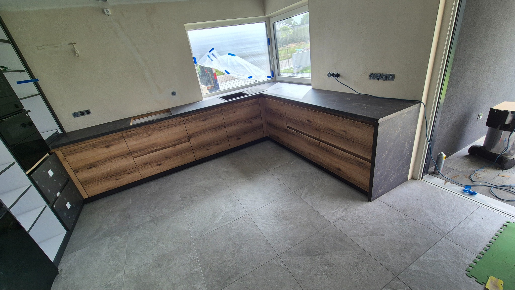 Kątowe ujęcie na zamontowaną, drewnopodobną zabudowę kuchenną z ciemnym blatem, widoczną na tle okna i szarej podłogi. Częściowo widoczny regał i odkurzacz.