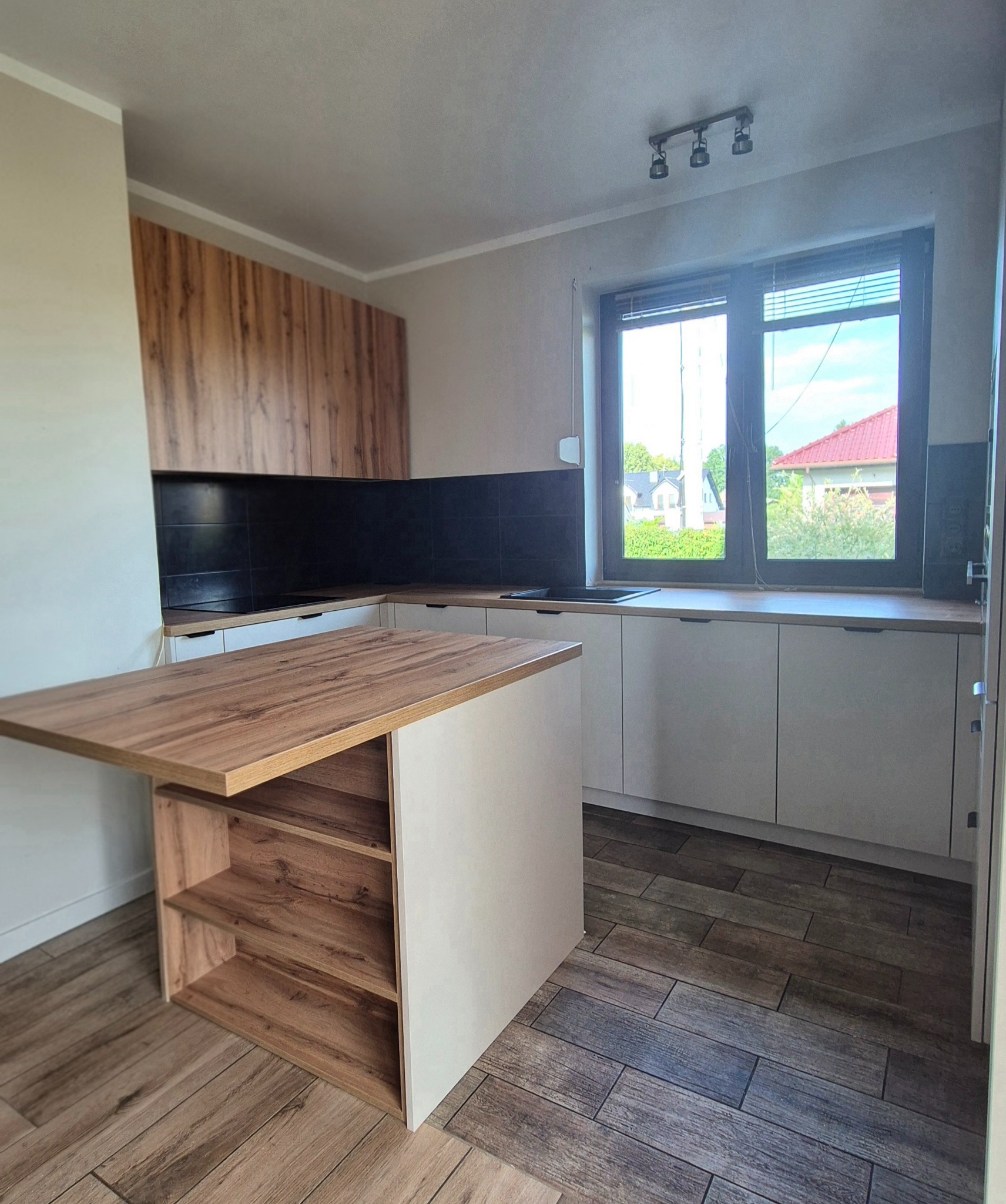 Jasna kuchnia z drewnianymi akcentami: wyspa z półkami, szafki pod oknem, drewniane panele na ścianie. Widok na domy przez okno.