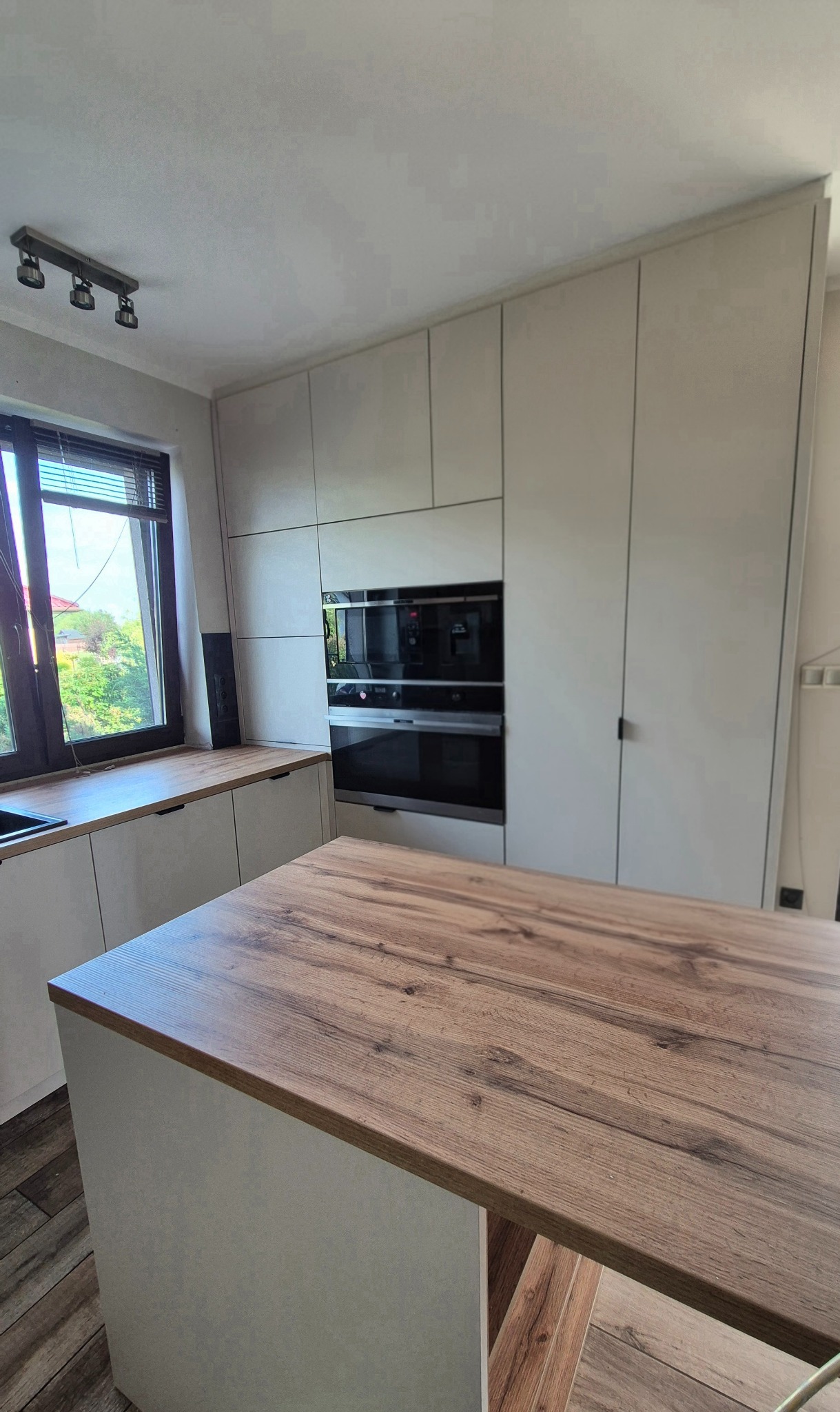 Jasna kuchnia z drewnianym blatem wyspy, zabudowa pod sufit z piekarnikiem i szafkami. Widok z bliska na blat wyspy i okno z żaluzjami.