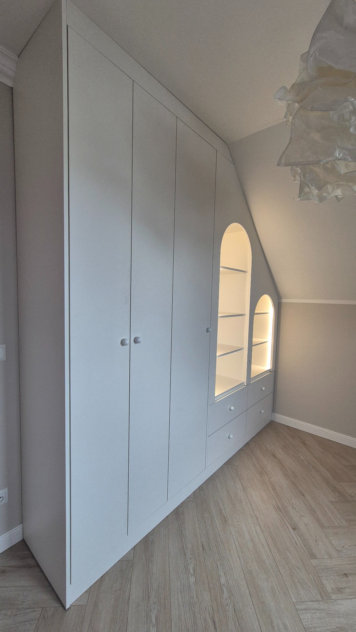Biała szafa wnękowa z oświetlonymi półkami w kształcie łuków, dopasowana do skosu poddasza, z drewnianą podłogą w jodełkę.