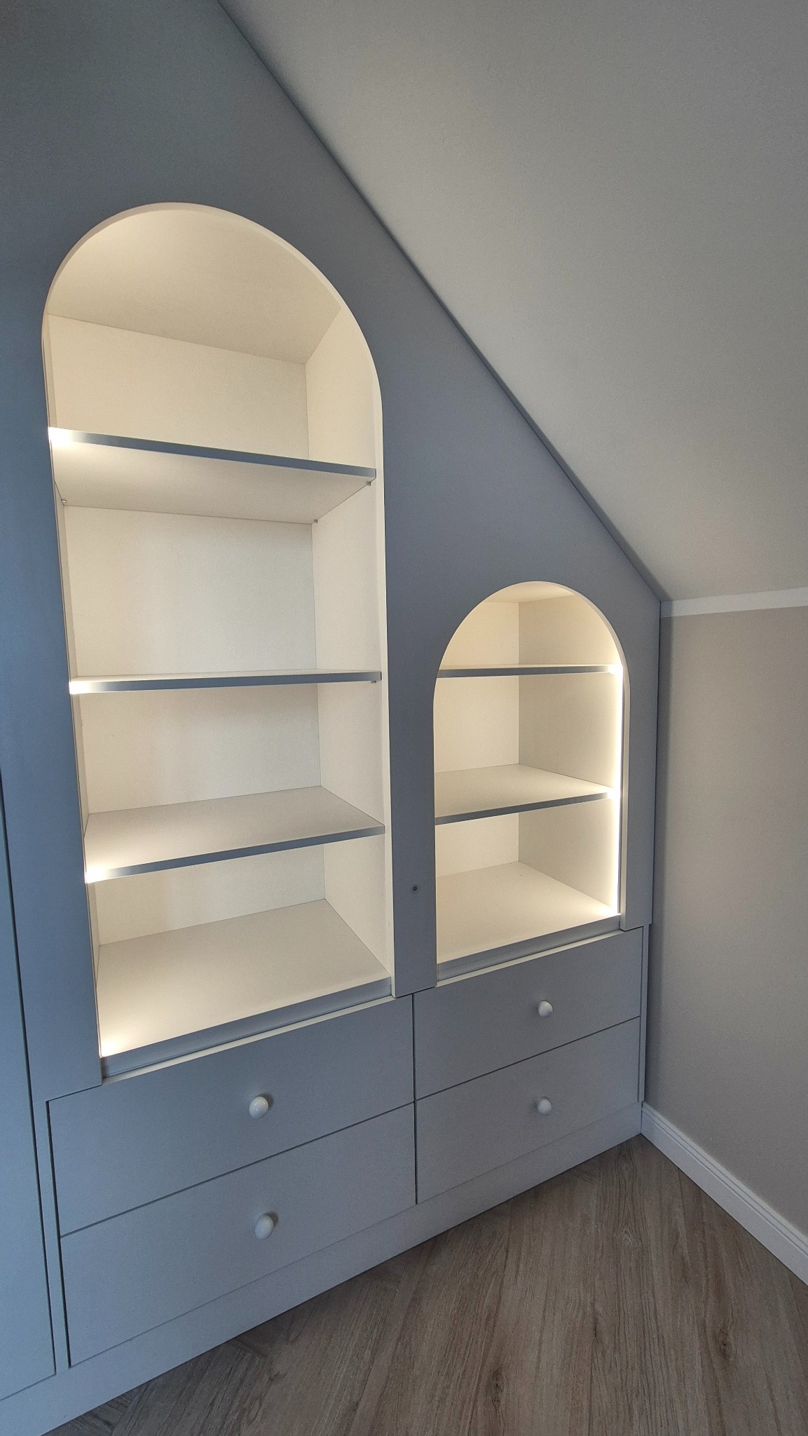 Niebieska szafa wnękowa z oświetleniem LED, wkomponowana w skos dachu, z półkami w kształcie łuków i szufladami z okrągłymi uchwytami.