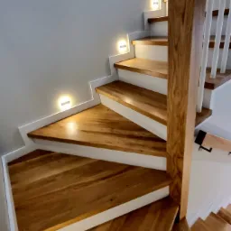 Drewniane schody z białymi podstopnicami i oświetleniem LED w ścianie, widok z dołu, ukazujący naturalny rysunek drewna.