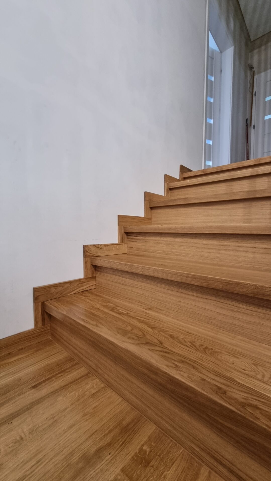 Detale drewnianych schodów z litego dębu, widoczne słoje drewna i precyzyjne wykończenie, minimalistyczne tło z białą ścianą i fragmentem okna.