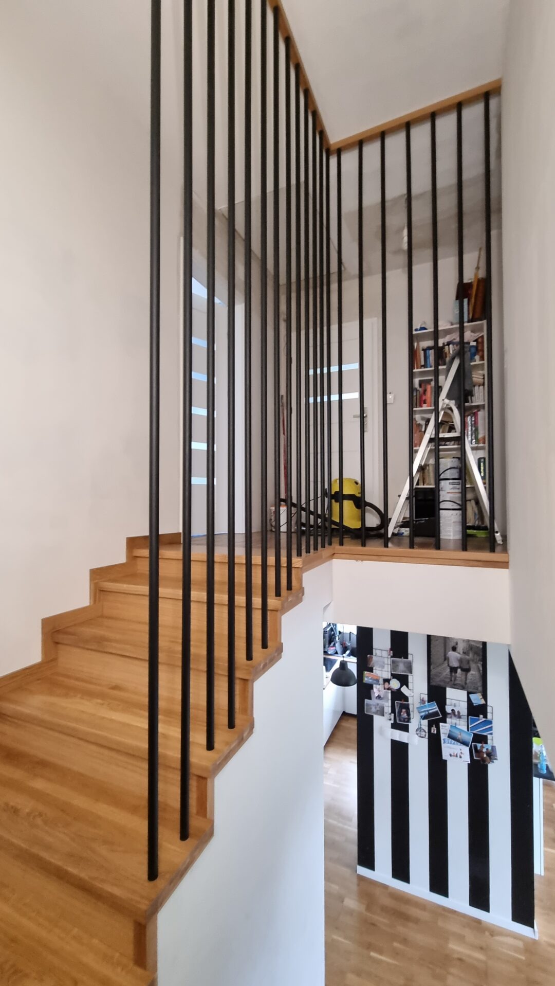 Drewniane schody z czarną, metalową balustradą w nowoczesnym wnętrzu z widocznym fragmentem kuchni i dekoracyjną ścianą w czarno-białe pasy.