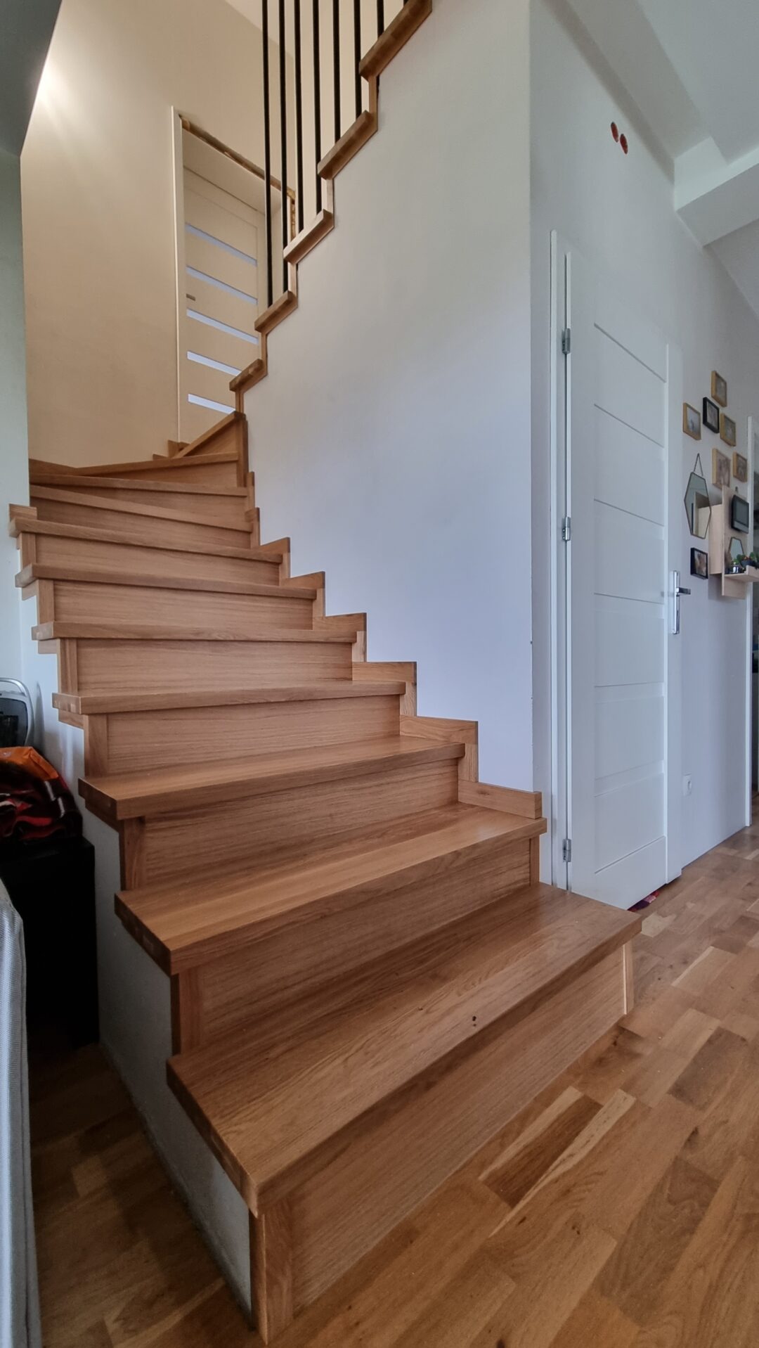 Drewniane schody z czarną balustradą w jasnym wnętrzu domu, widoczne drzwi wewnętrzne z przeszkleniami i fragment podłogi z parkietu.