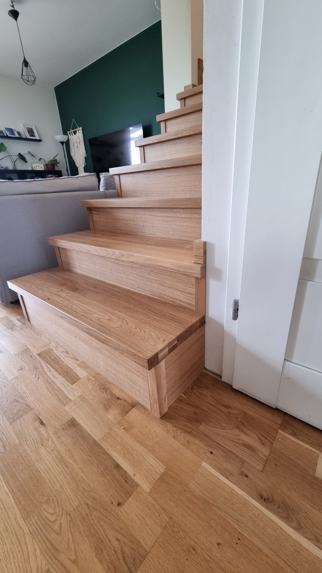 Drewniane schody dębowe z widocznymi połączeniami czopowymi, prowadzące w górę, z fragmentem salonu w tle i białą ścianą z drzwiami po prawej stronie.