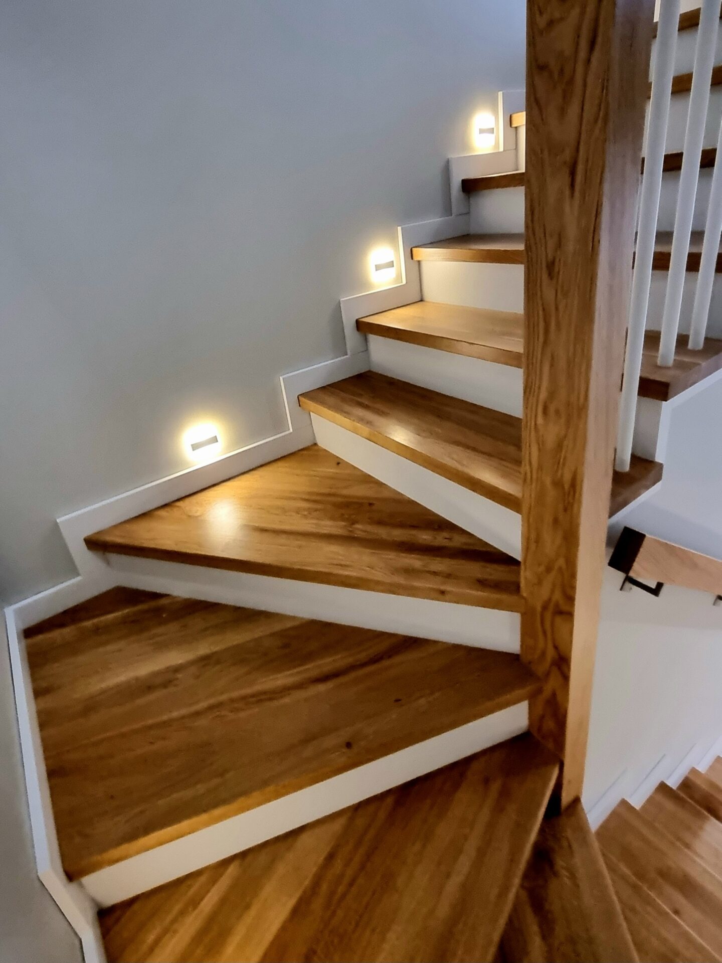 Drewniane schody z białymi podstopnicami i oświetleniem LED w ścianie, widok z dołu, ukazujący naturalny rysunek drewna.