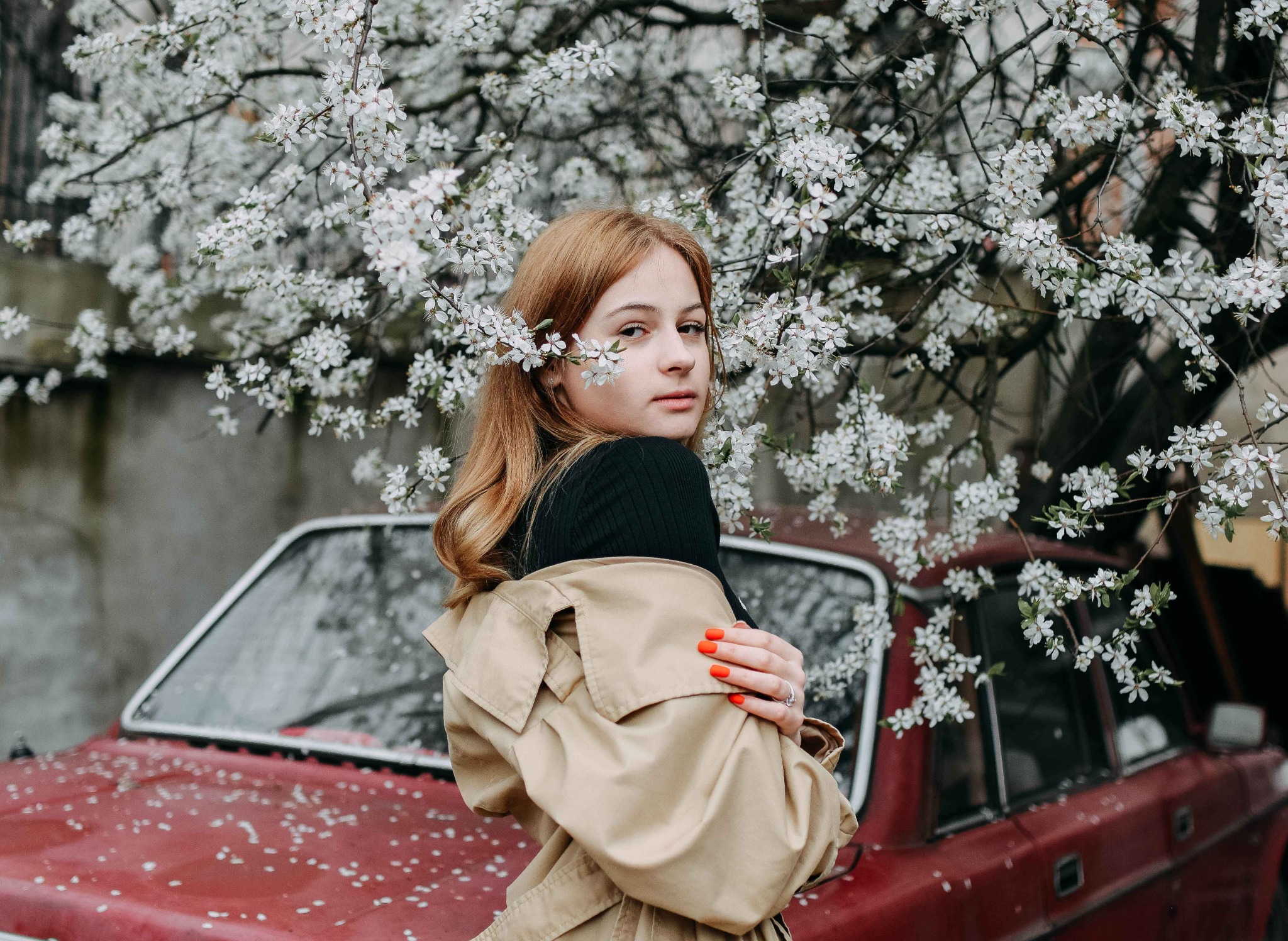 Portret kobiety w beżowym płaszczu, otoczonej kwitnącym drzewem i czerwonym samochodem w tle, z kwiatami we włosach i pomarańczowymi paznokciami.