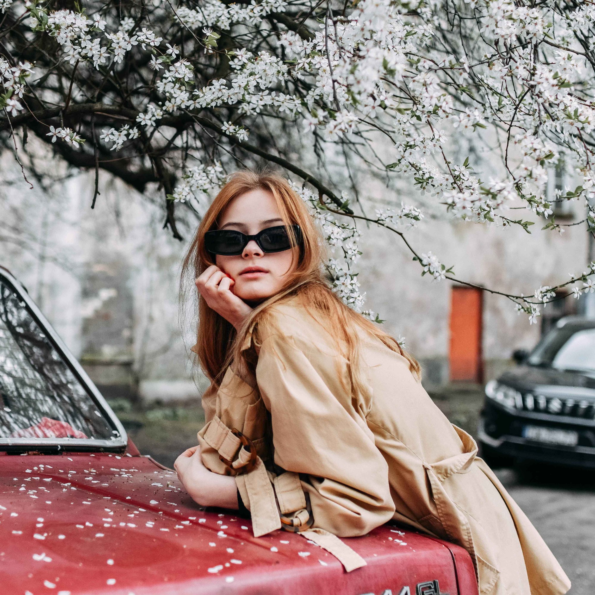 Młoda kobieta w okularach przeciwsłonecznych i beżowym płaszczu opiera się o czerwoną maskę starego samochodu, na której leżą opadłe białe kwiaty, w tle kwitnące drzewo i budynek.