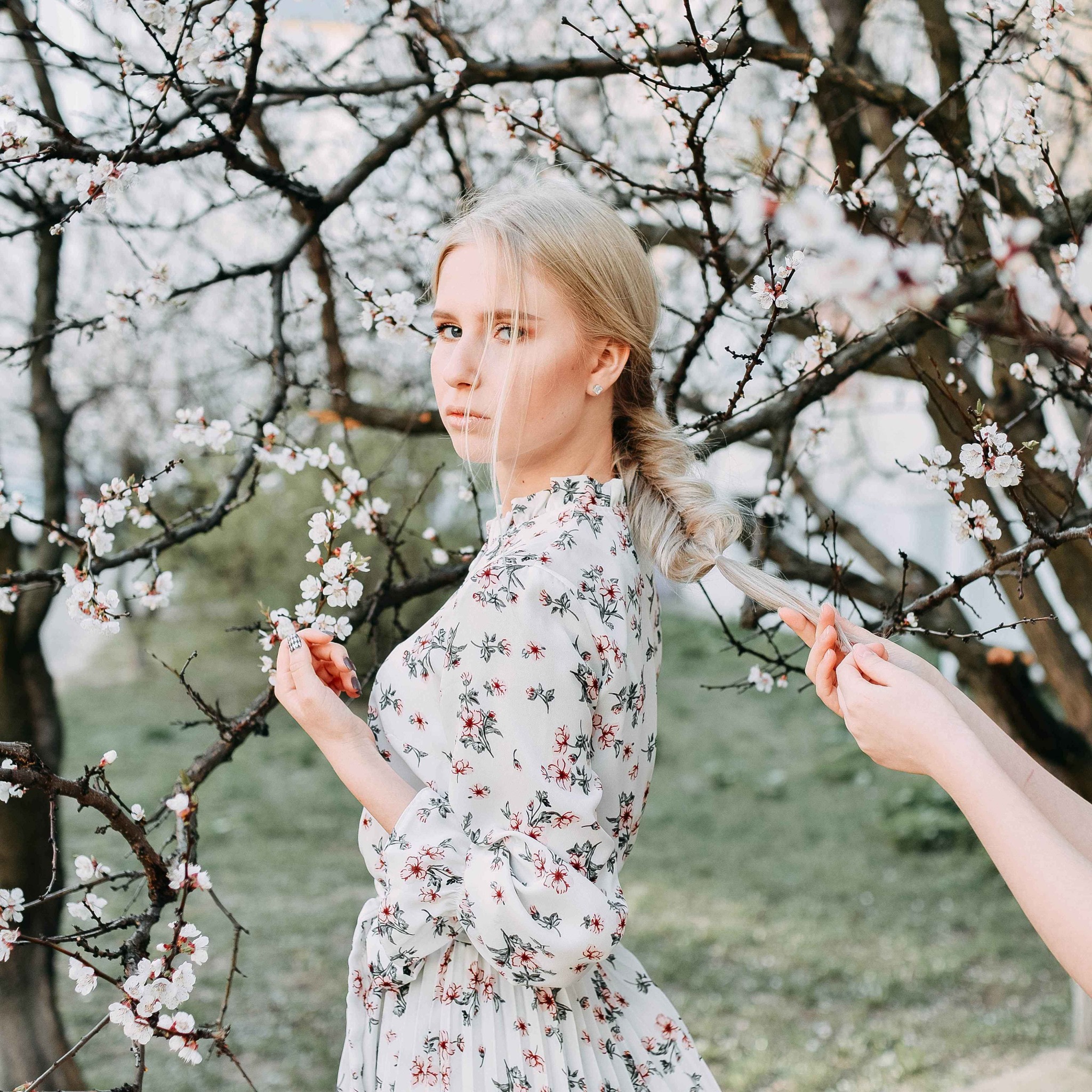 Portret młodej kobiety z blond włosami w kwiecistej sukience, stojącej na tle kwitnącego drzewa, z dłońmi stylizującymi warkocz