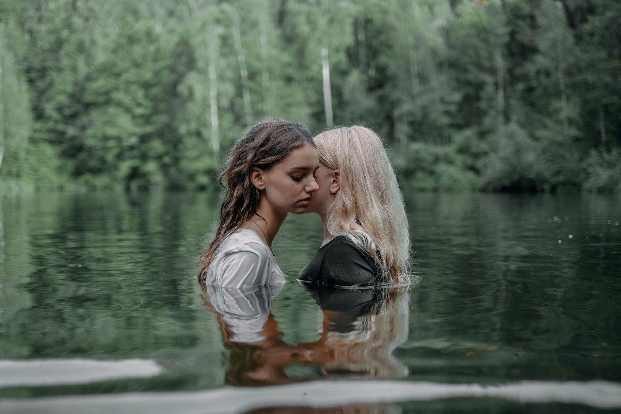 Dwie kobiety zanurzone do ramion w wodzie jeziora, z bliska, z odbiciem w tafli wody, na tle zielonego lasu.