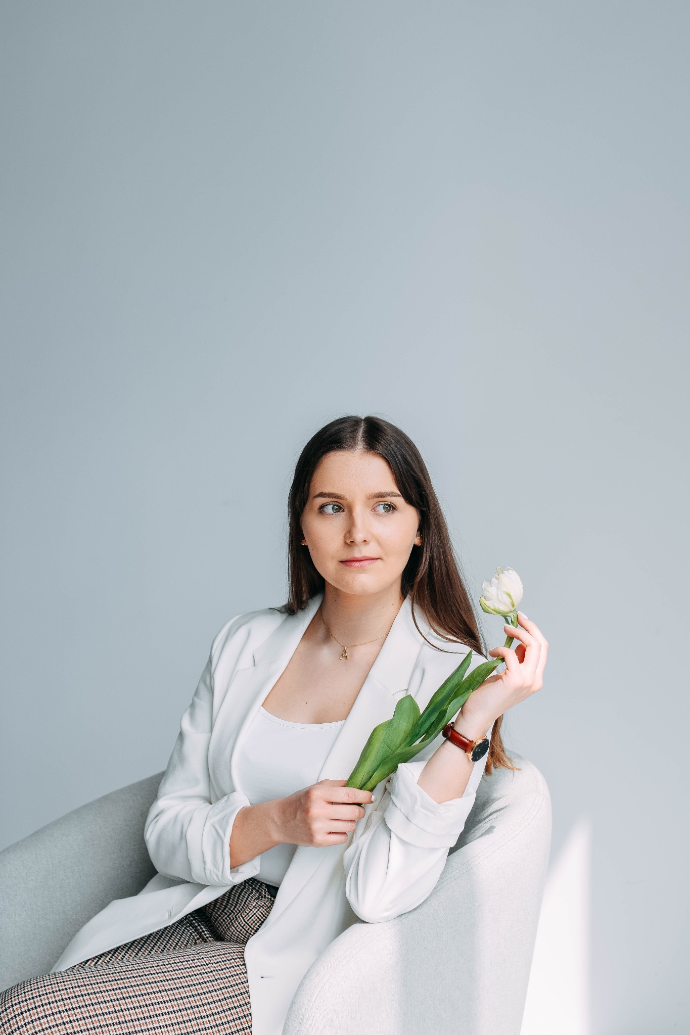 Kobieta w białym żakiecie siedzi w fotelu, trzymając białego tulipana z zielonymi liśćmi, spoglądając w bok.