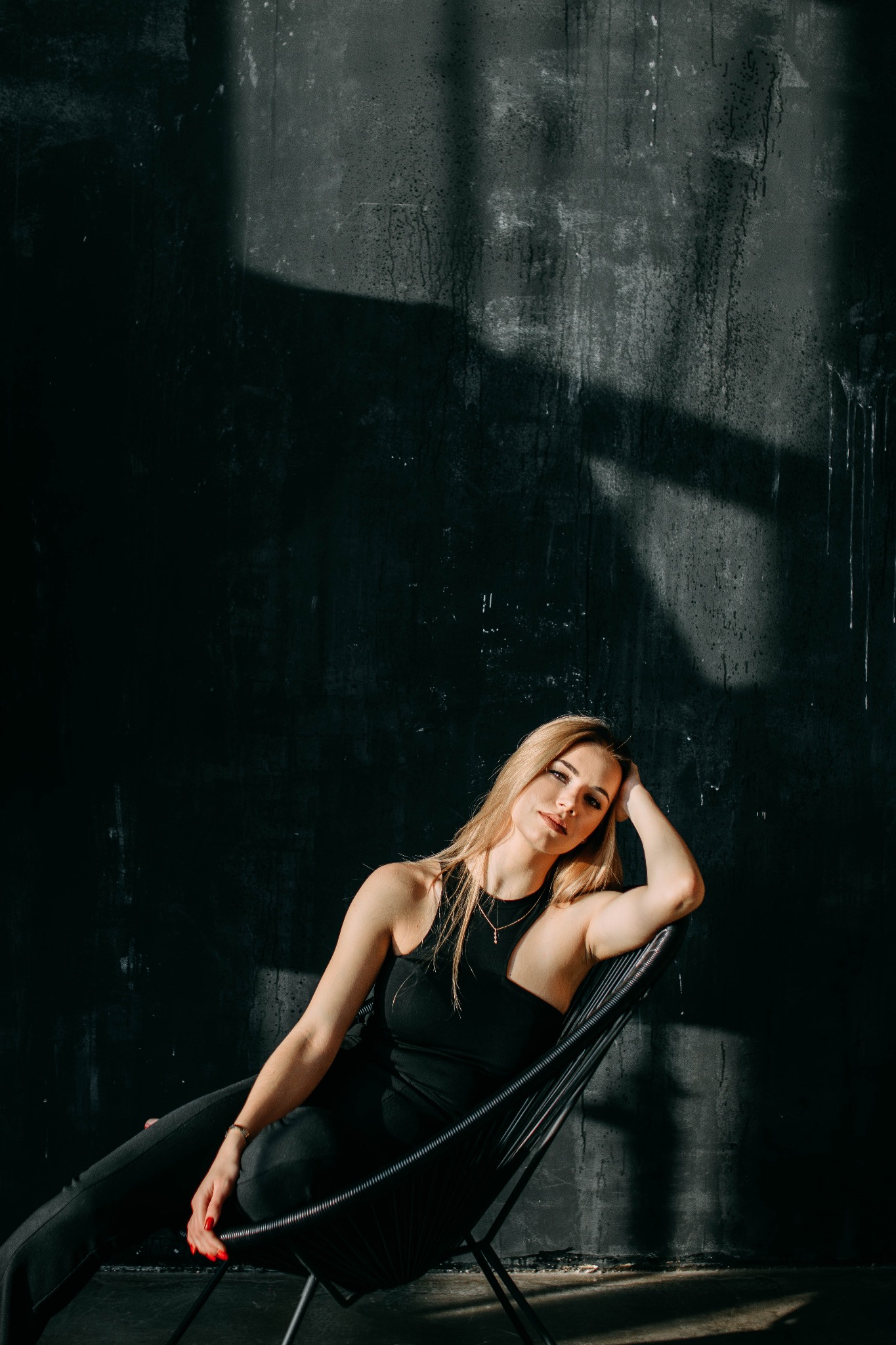 Portret kobiety z blond włosami, ubranej na czarno, siedzącej w czarnym, designerskim fotelu na tle ciemnej, teksturowanej ściany z plamami światła