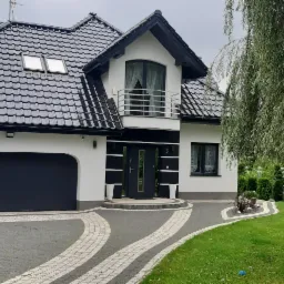 Nowoczesny dom jednorodzinny z czarną bramą garażową segmentową i drzwiami wejściowymi w kolorze antracytu, widok z zewnątrz na podjazd z kostki brukowej.