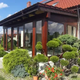 Zadaszony taras z przesuwnymi drzwiami w Brzegu Dolnym, otoczony ozdobną roślinnością i brukowanym chodnikiem, widok z zewnątrz w słoneczny dzień.