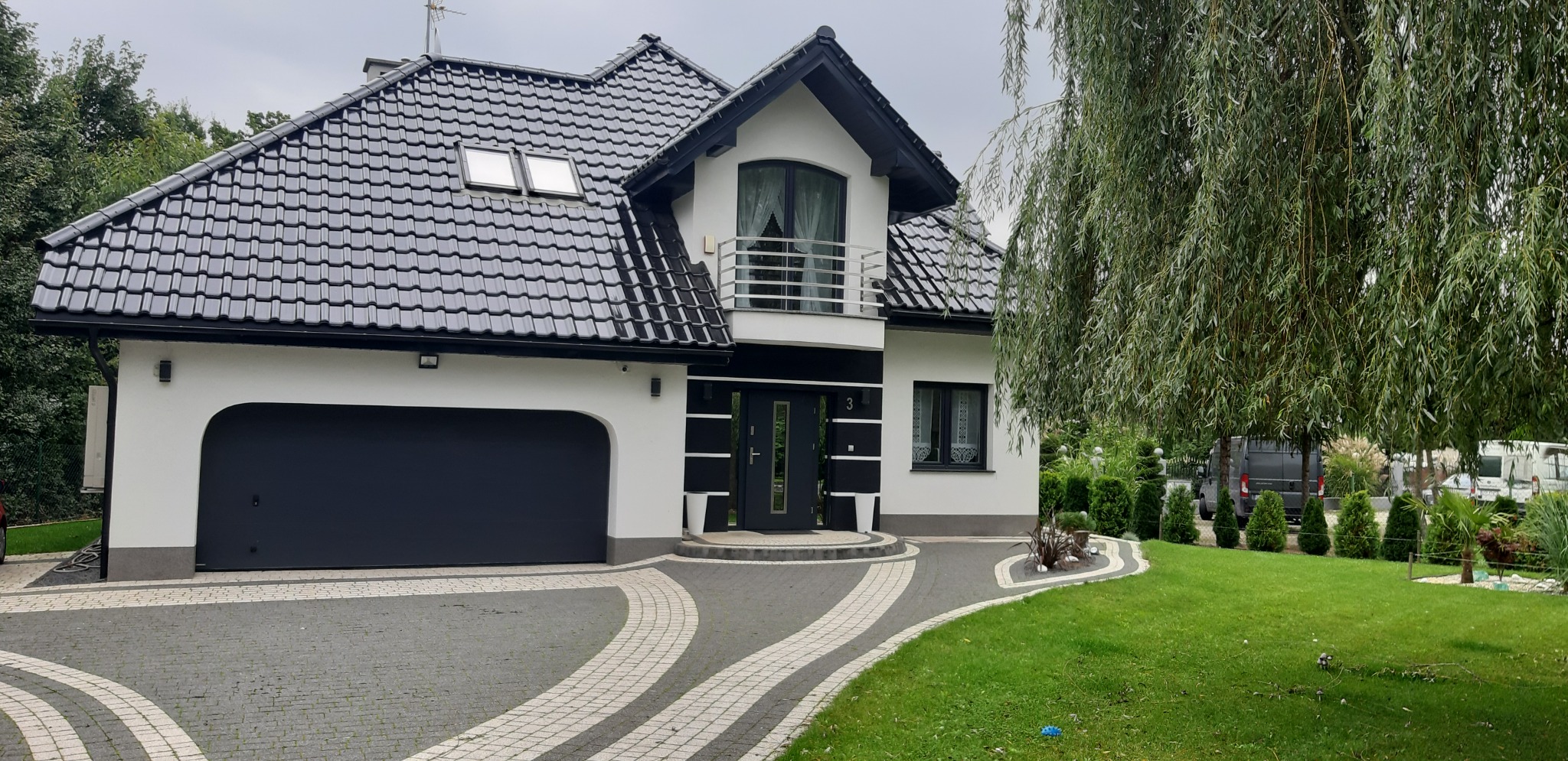 Nowoczesny dom jednorodzinny z czarną bramą garażową segmentową i drzwiami wejściowymi w kolorze antracytu, widok z zewnątrz na podjazd z kostki brukowej.