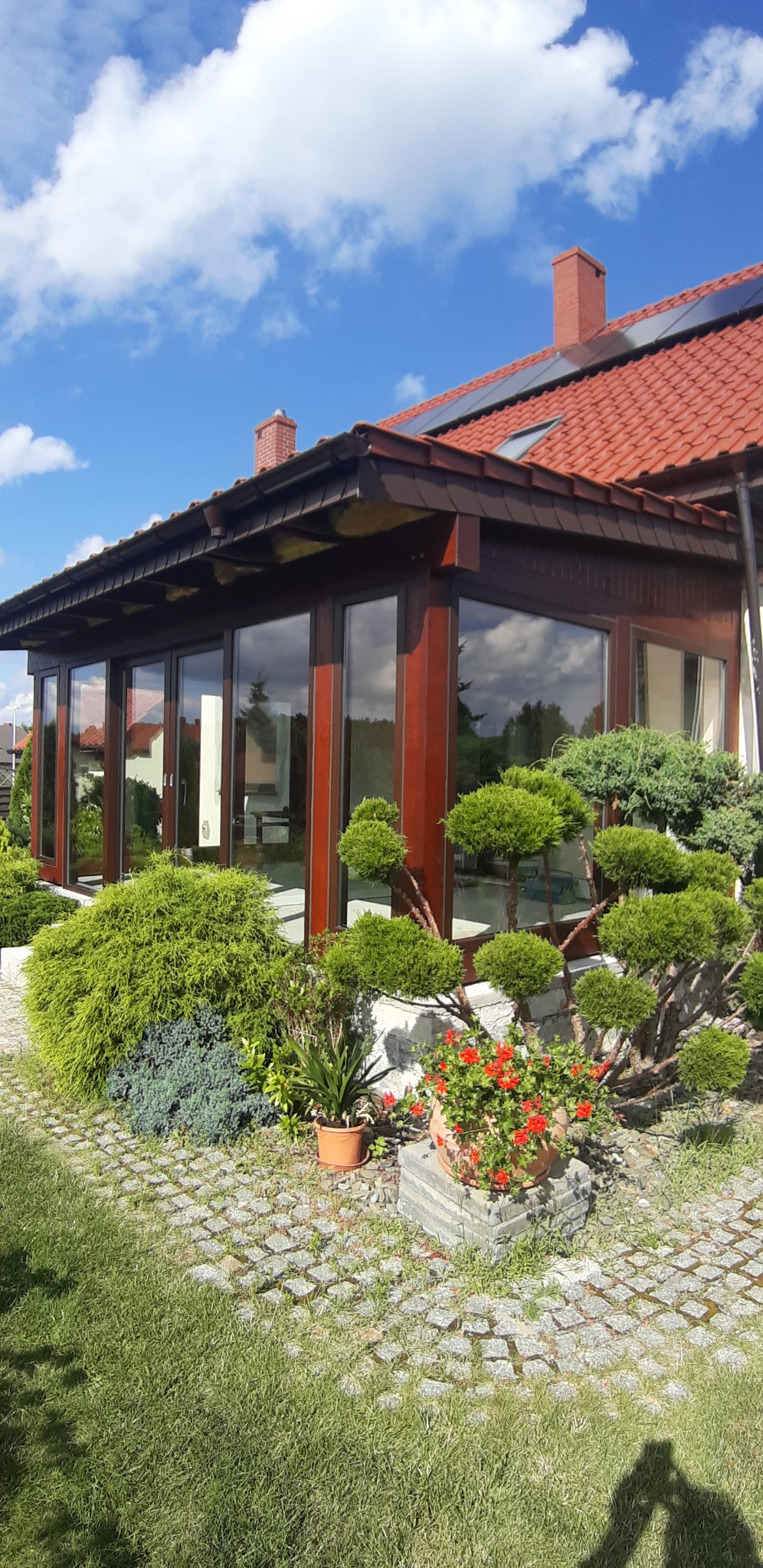 Zadaszony taras z przesuwnymi drzwiami w Brzegu Dolnym, otoczony ozdobną roślinnością i brukowanym chodnikiem, widok z zewnątrz w słoneczny dzień.