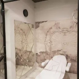 Pokój z lustrem w romby i dekoracyjną tapetą w stylu vintage, z białym fotelem kosmetycznym przykrytym pokrowcem.