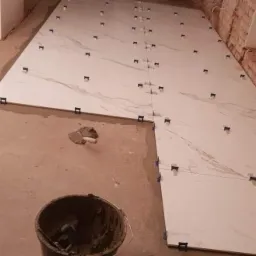 Układanie białych, marmurkowych płytek podłogowych z użyciem klipsów poziomujących, widoczny mur z czerwonej cegły w tle.