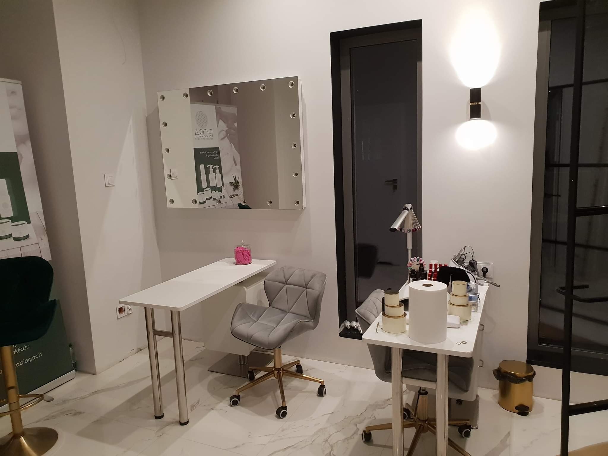 Nowoczesne wnętrze salonu kosmetycznego z dwoma stanowiskami do manicure: białe biurka na metalowych nogach, szare krzesła na złotych kółkach, lustro z oświetleniem i lampa na ścianie.