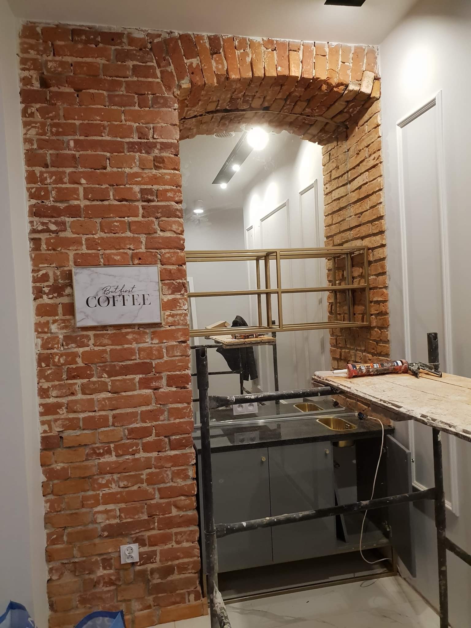 Wnętrze w trakcie prac wykończeniowych: widoczna ceglana kolumna z łukiem, pod nią szara szafka z dwoma złotymi zlewami, rusztowanie i narzędzia, na cegle wisi tabliczka z napisem 'But first coffee'.