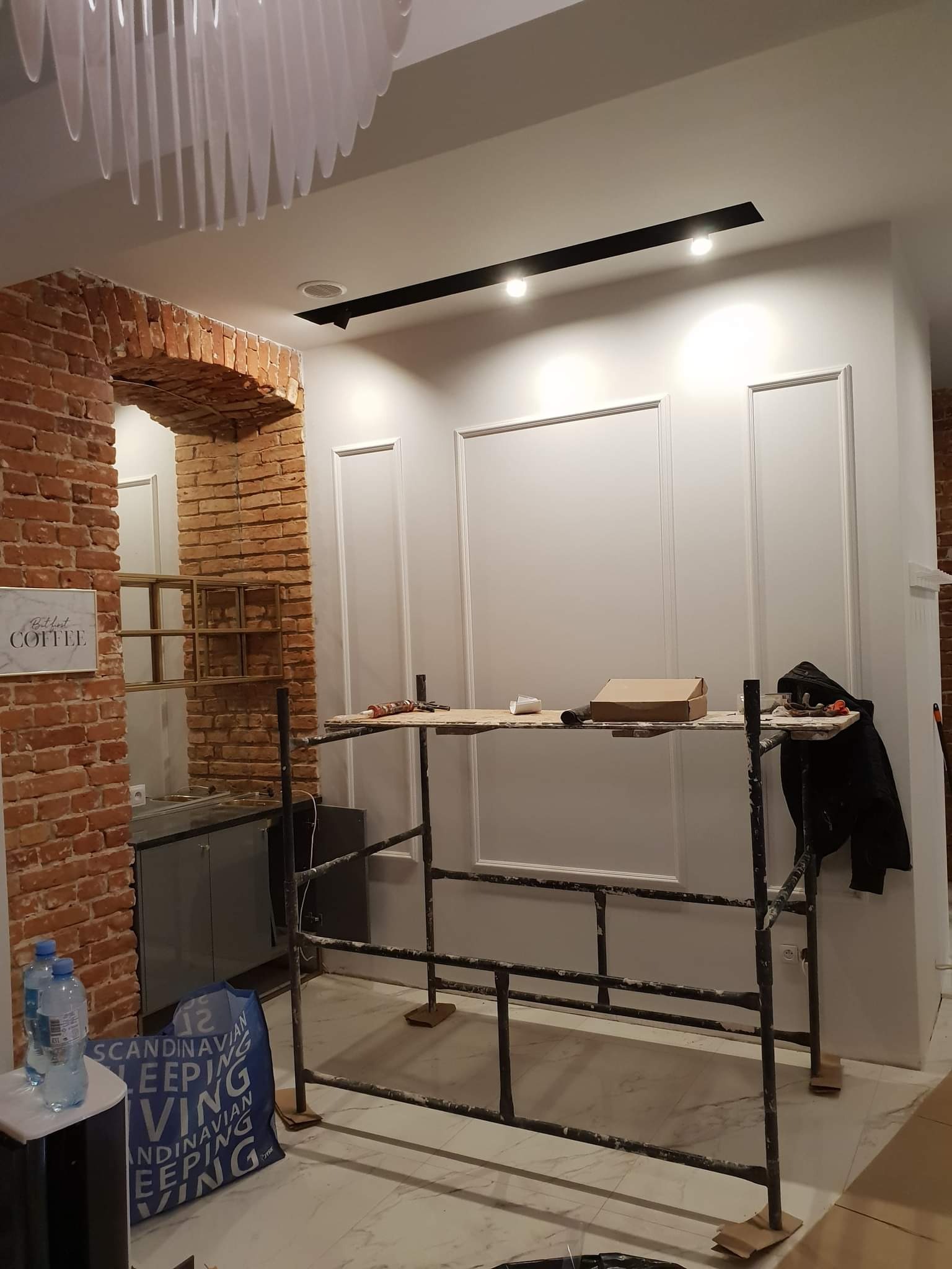 Wnętrze w trakcie wykańczania z widocznym rusztowaniem, ceglanym łukiem i panelami ściennymi w stylu klasycznym. Na rusztowaniu narzędzia i karton, w tle widoczna instalacja oświetleniowa.