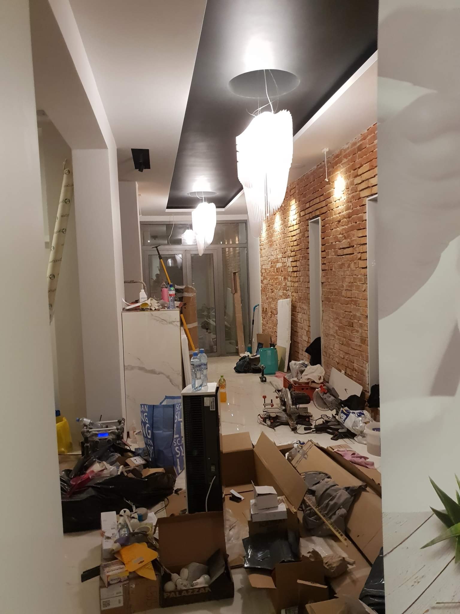 Wnętrze w trakcie prac wykończeniowych: odsłonięta cegła na ścianie, nowoczesne lampy wiszące, widoczne narzędzia i materiały budowlane w nieładzie.