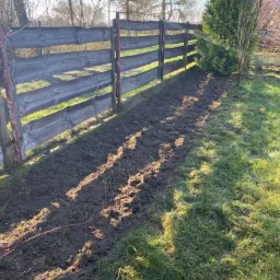 Przygotowany pas ziemi wzdłuż drewnianego płotu, gotowy do sadzenia, z widoczną tują przy płocie i trawnikiem po prawej stronie, w słoneczny dzień.