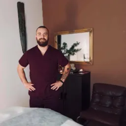 Uśmiechnięty mężczyzna w bordowym uniformie medycznym, z tatuażem na ramieniu, stoi w gabinecie z kozetką przykrytą jednorazowym prześcieradłem, na ścianie wisi drewniana maska i lustro w złotej...