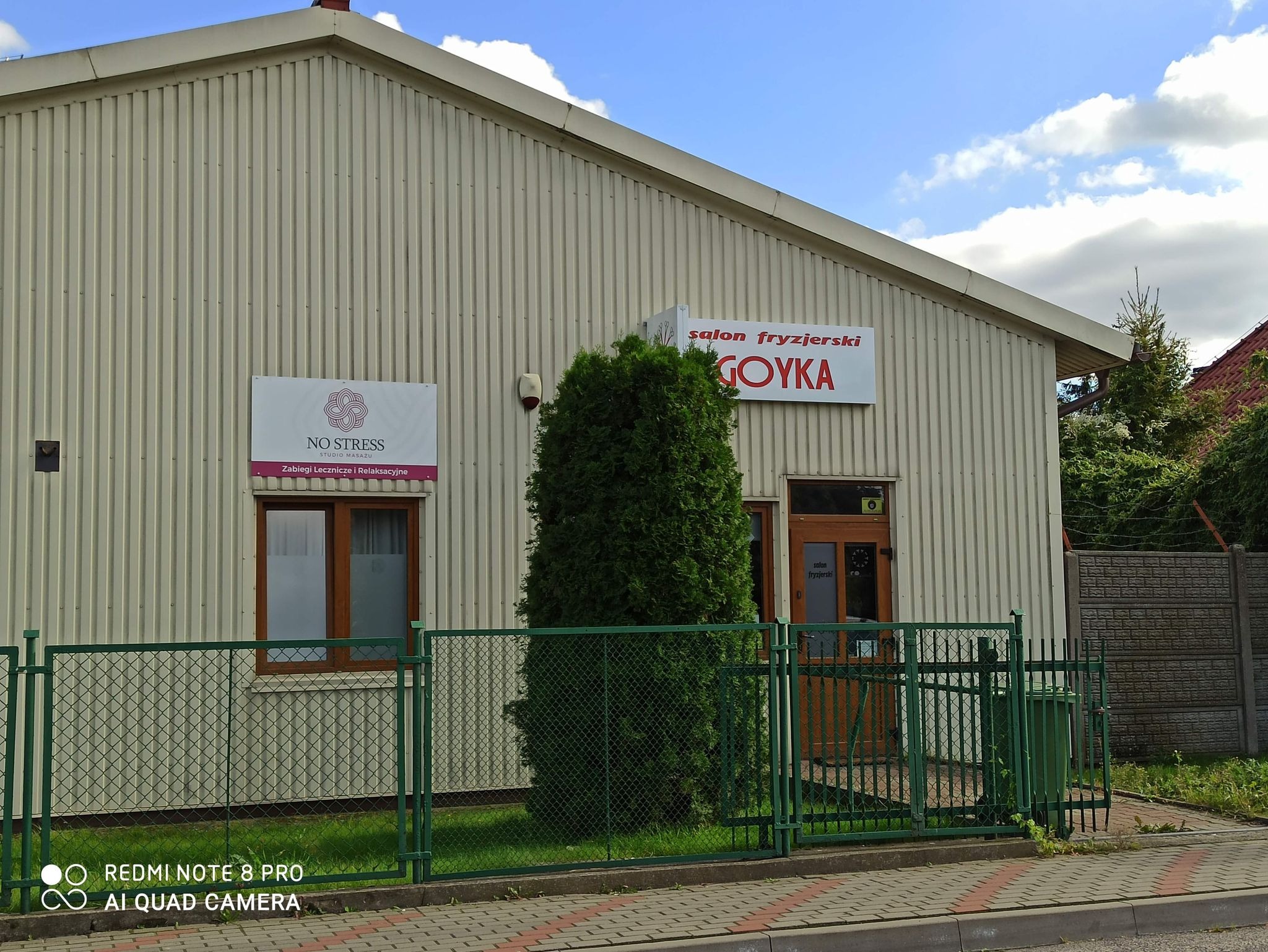 Budynek z dwoma szyldami: 'NO STRESS Studio Masażu Zabiegi Lecznicze i Relaksacyjne' oraz 'Salon Fryzjerski GOYKA', otoczony zielonym ogrodzeniem i tujami.