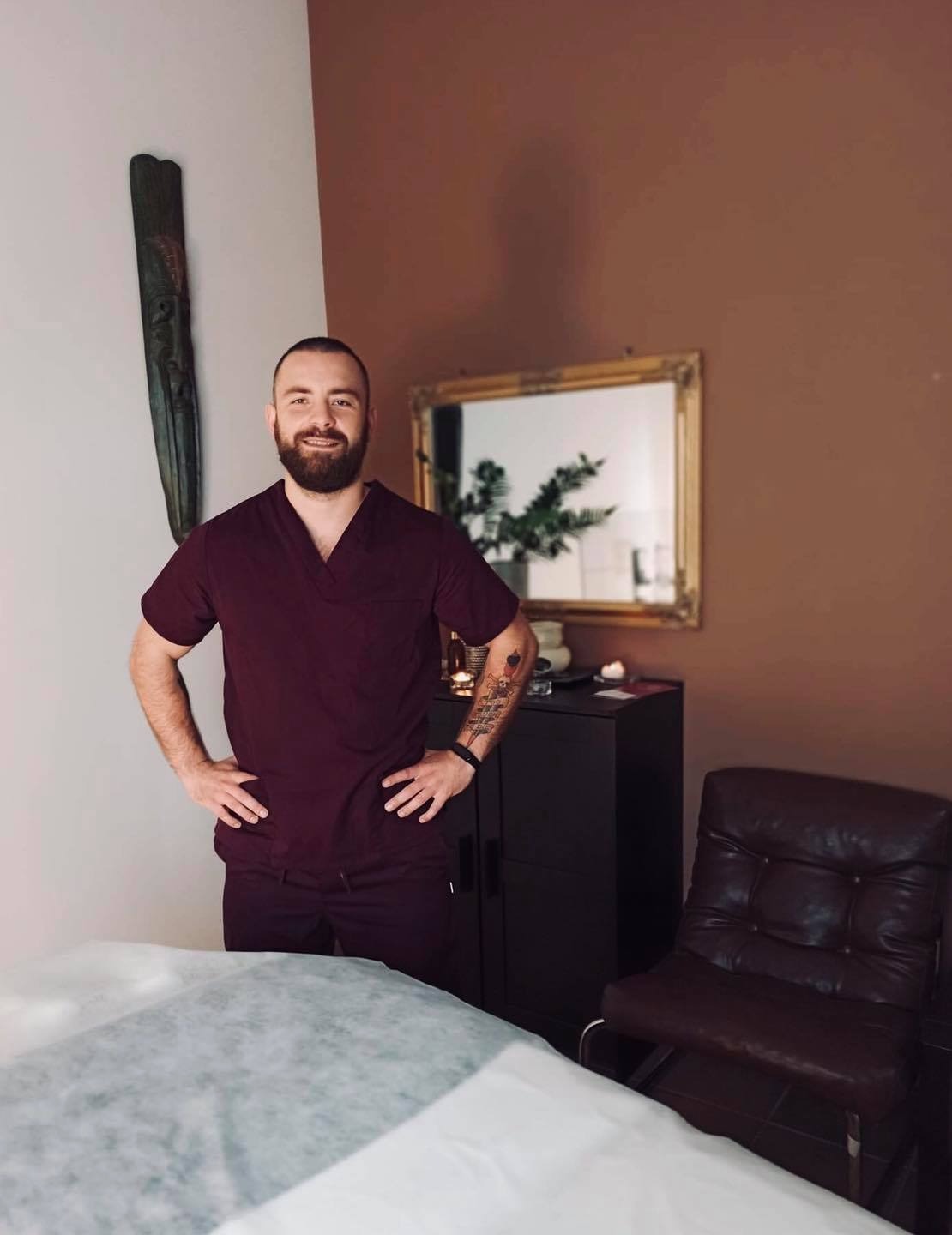 Uśmiechnięty mężczyzna w bordowym uniformie medycznym, z tatuażem na ramieniu, stoi w gabinecie z kozetką przykrytą jednorazowym prześcieradłem, na ścianie wisi drewniana maska i lustro w złotej...