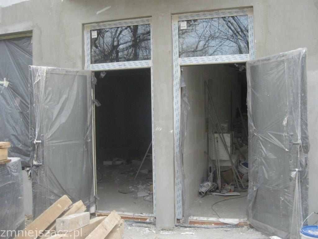 Dwa wejścia z zamontowaną stolarką okienną PCV, zabezpieczone folią ochronną, w trakcie prac wykończeniowych na surowym murze.