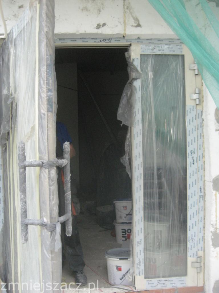 Montaż drzwi balkonowych PCV w trakcie prac wykończeniowych; zabezpieczone folią, widoczne wiaderka z farbą i narzędzia.