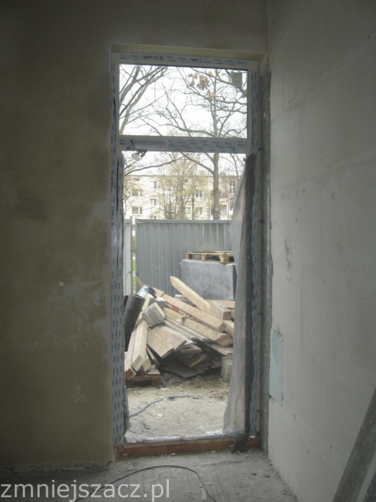 Surowy montaż ramy okiennej PCV w murze, widoczne drewniane deski za oknem i surowe ściany w trakcie wykończenia.