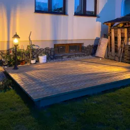 Oświetlony drewniany taras przydomowy z widocznymi lampami ogrodowymi, roślinami w donicach i składem drewna w tle, sfotografowany wieczorem.