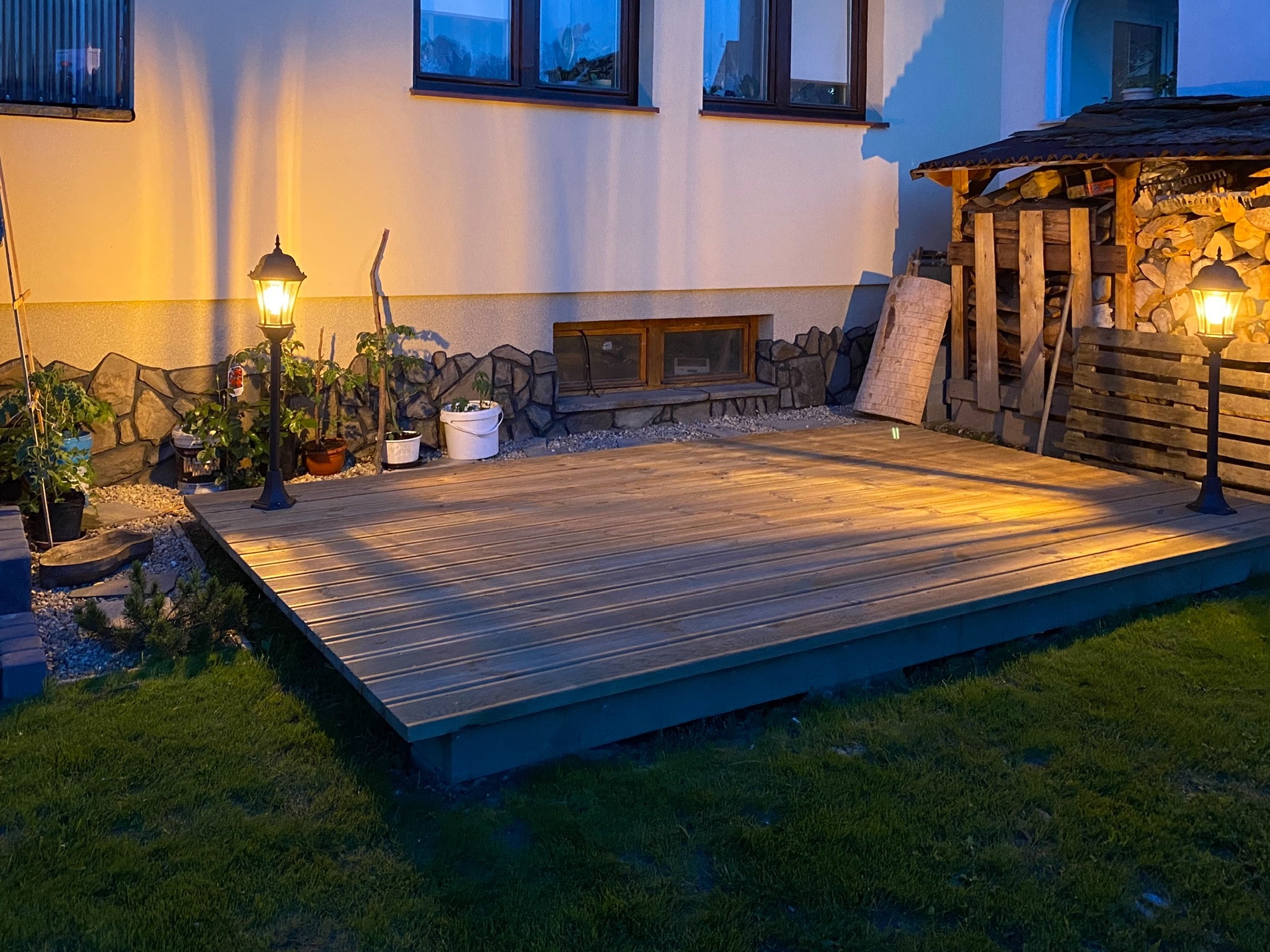 Oświetlony drewniany taras przydomowy z widocznymi lampami ogrodowymi, roślinami w donicach i składem drewna w tle, sfotografowany wieczorem.