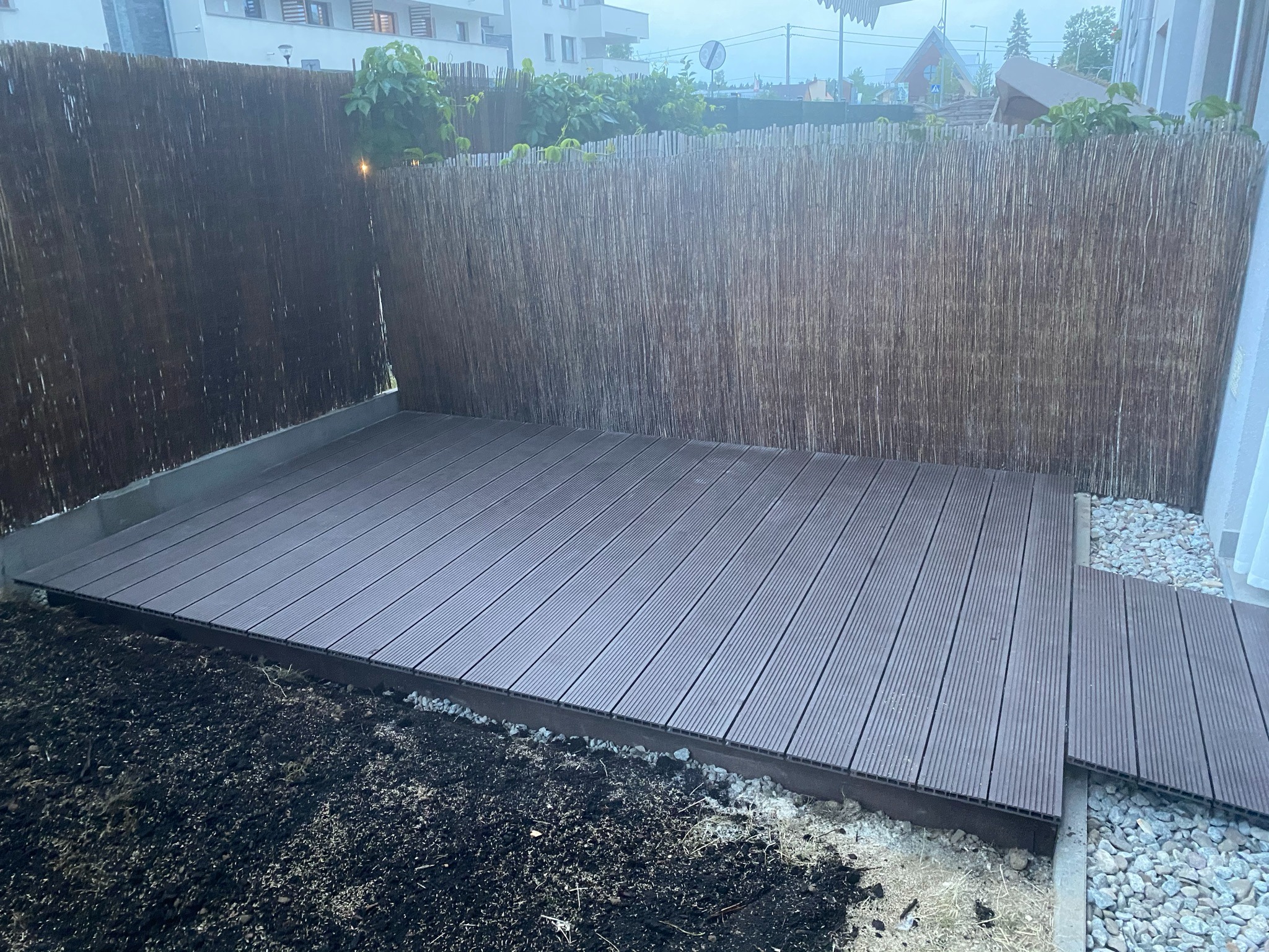 Nowo ułożony, brązowy taras kompozytowy z ryflowaną powierzchnią, otoczony ziemią i kamieniami, przylegający do trzcinowego ogrodzenia i budynku mieszkalnego w tle.