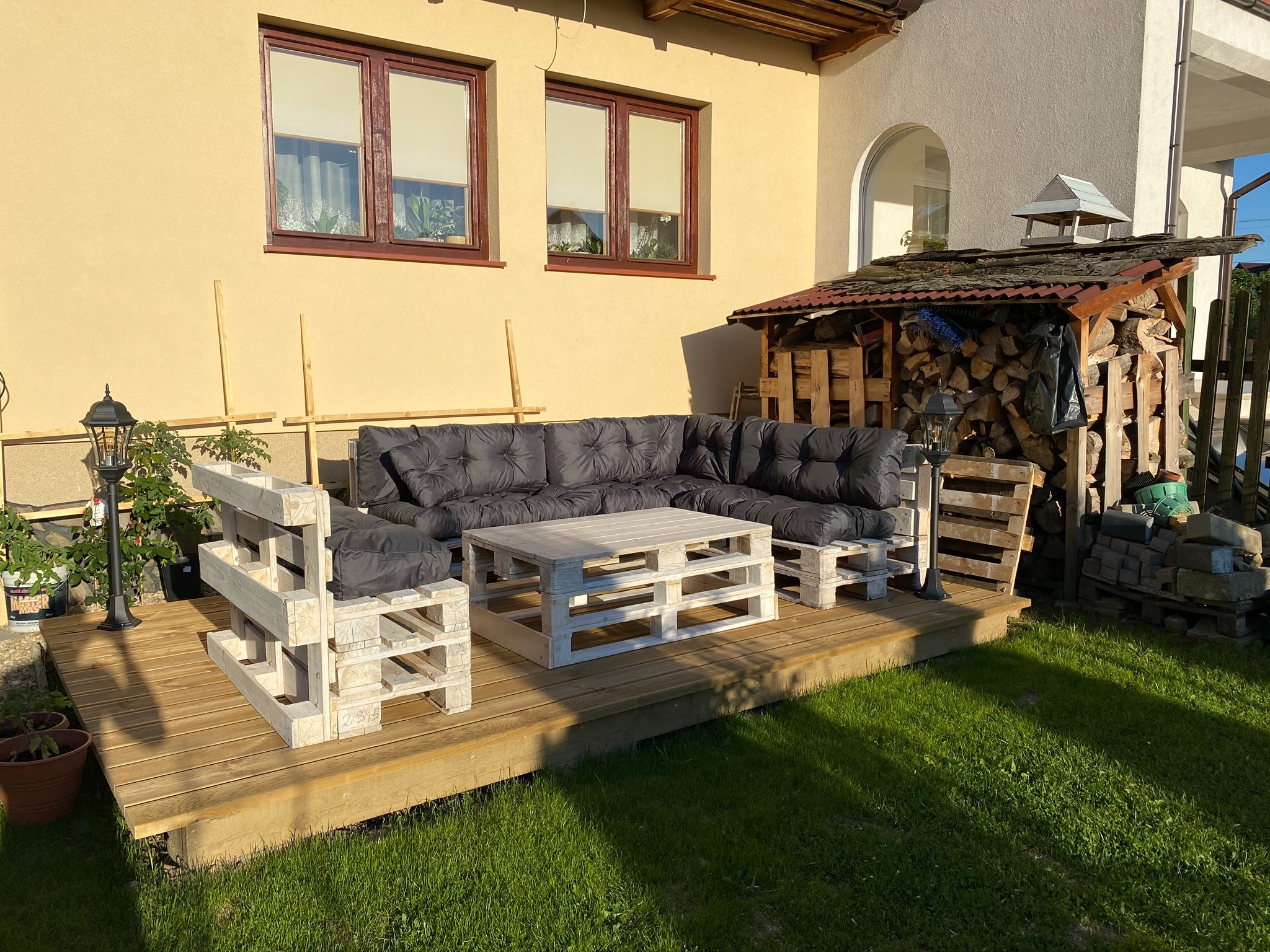 Drewniany taras z meblami ogrodowymi wykonanymi z palet pomalowanych na biało, z szarymi poduszkami, obok budynku z beżową elewacją i stertą drewna pod zadaszeniem.