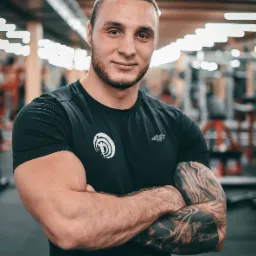 Uśmiechnięty mężczyzna z brodą i tatuażami na ramieniu, ubrany w czarną koszulkę z logo, na tle siłowni z widocznymi maszynami do ćwiczeń.