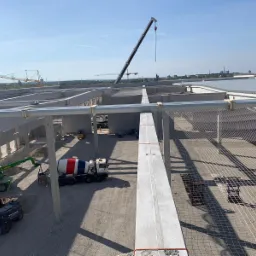 Budowa hali: widok z góry na betonowe słupy konstrukcyjne, żuraw, betoniarkę z czerwono-biało-niebieskim wzorem, siatkę zabezpieczającą i maszyny budowlane w słoneczny dzień.