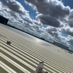 Jasna, blaszana konstrukcja dachu przemysłowego z widocznymi elementami mocującymi, na tle błękitnego nieba z chmurami, z elementami opakowania Wurth na pierwszym planie.